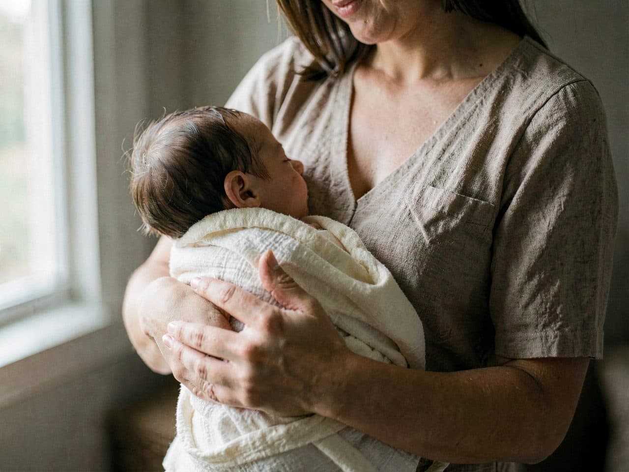 A parent holding a sleeping baby by a window in soft morning light
